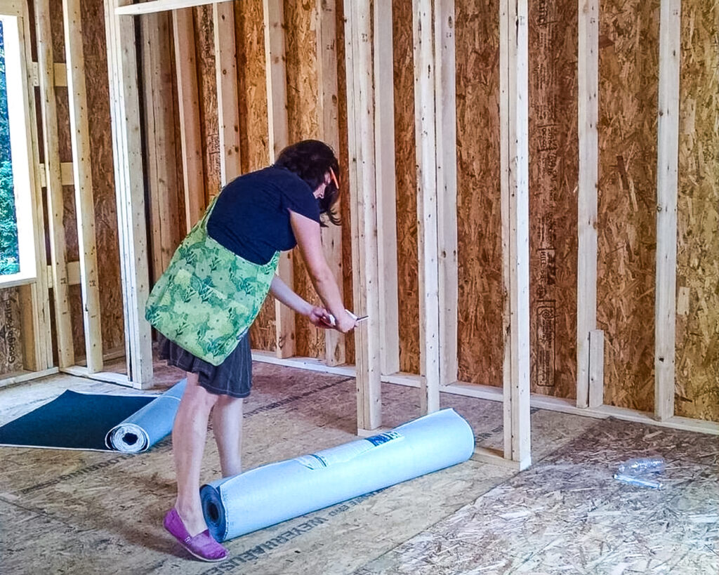 Leesa, a DFC-LMA architect, is at a construction site examining a roll of material on an unfinished floor. The exterior and interior wall are just wood framing and you can see an open window to the left